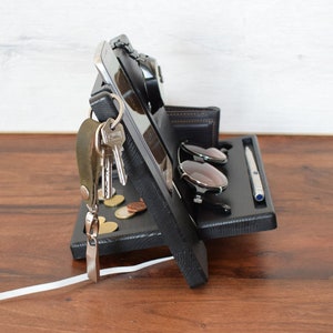 Puede incluir: Organizador de madera negro con compartimentos para un tel&eacute;fono, un reloj, llaves, gafas de sol y un bol&iacute;grafo. El organizador tiene una superficie texturizada y una peque&ntilde;a cartera de cuero.