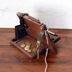 May include: Wooden organiser with compartments for a phone, watch, keys, and wallet. It has a tray for loose change and a slot for charging cables.