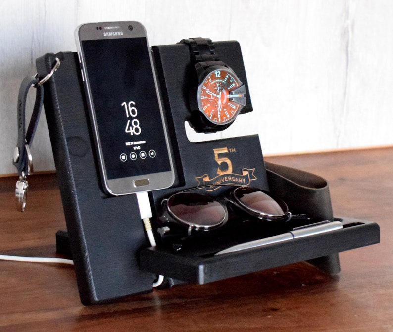 Puede incluir: Organizador de madera negro con ranuras para un tel&eacute;fono, un reloj, gafas de sol y un bol&iacute;grafo. El organizador tiene una inscripci&oacute;n de 5&ordm; aniversario y un gancho para llaves.
