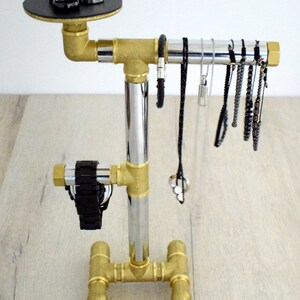 May include: A gold metal jewelry stand with a black circular top for rings. The stand has a chrome vertical pipe and a horizontal pipe for hanging necklaces and bracelets. The stand has a base with four legs.