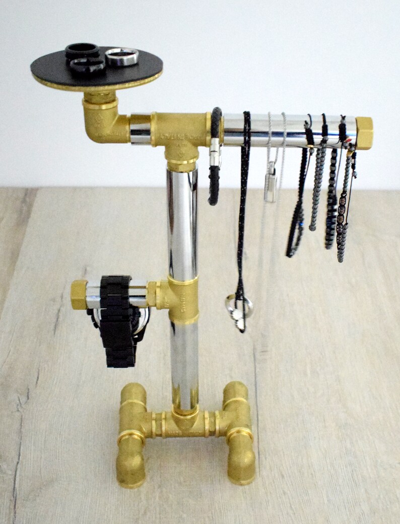 May include: A gold metal jewelry stand with a black circular top for rings. The stand has a chrome vertical pipe and a horizontal pipe for hanging necklaces and bracelets. The stand has a base with four legs.