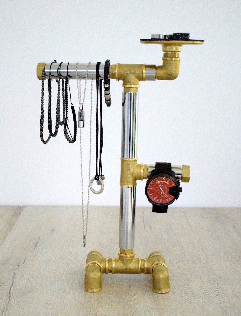 May include: A gold metal jewelry stand with a silver pipe and a watch holder. The stand has a round top and three legs. The stand is decorated with a watch and several necklaces and bracelets.