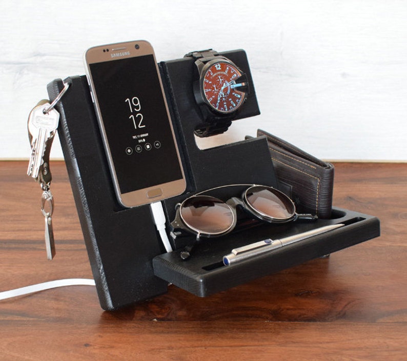 Puede incluir: Organizador de escritorio negro con compartimentos para un tel&eacute;fono, un reloj, gafas de sol y un bol&iacute;grafo. El organizador tiene un gancho para llaves y un cable de carga.