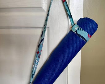 May include: A rolled royal blue yoga mat secured with a light blue fabric strap featuring a floral pattern in red, white, and dark blue. The strap includes two light-colored wooden rings and hangs from a dark-colored door handle.