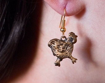 May include: Gold dangle earrings featuring a small, detailed bronze chicken charm.