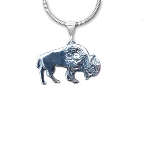 May include: A silver-colored pendant necklace featuring a detailed bison charm. The pendant is attached to a silver chain. The bison is sculpted with textured fur and a prominent head, showcasing the animal's form. The necklace is displayed against a white background.