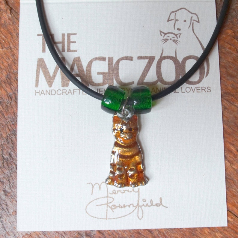 May include: A black cord necklace with a green glass bead charm and a silver charm of a sitting orange tabby cat. The necklace is on a white card with the text "The Magic Zoo Handcrafted Jewellery for Animal Lovers".