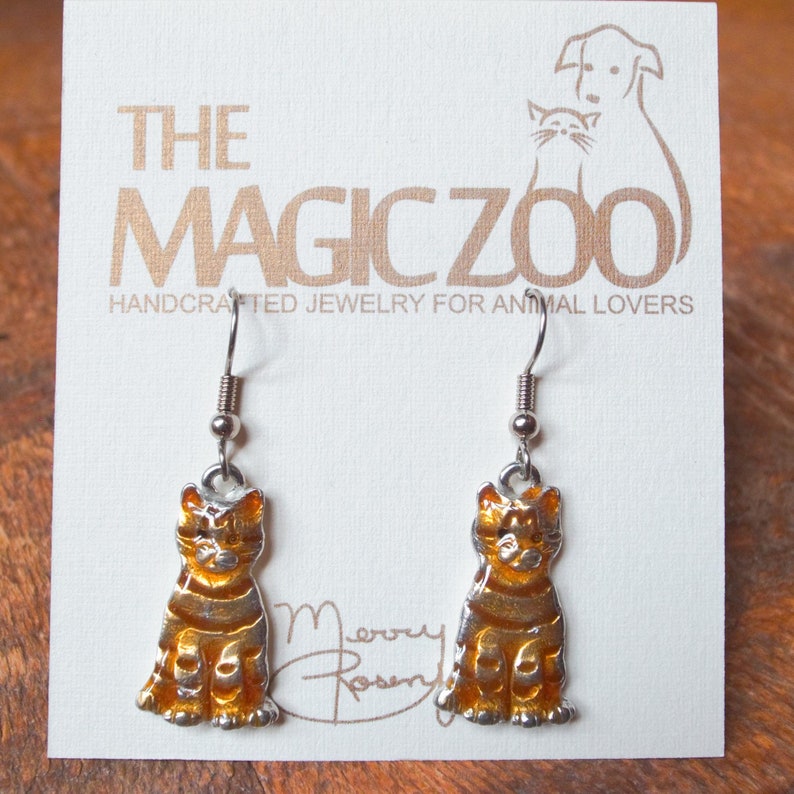 May include: A pair of silver cat earrings with orange and gold enamel. The earrings are on a white card with the text "The Magic Zoo Handcrafted Jewellery for Animal Lovers".
