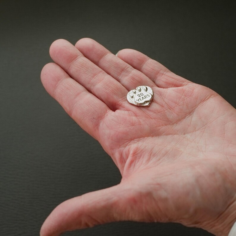 Puede incluir: Un amuleto de plata en forma de coraz&oacute;n con la inscripci&oacute;n "20 A&Ntilde;OS" en &eacute;l. El amuleto est&aacute; siendo sostenido en la mano de una persona.
