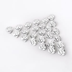 Puede incluir: Un conjunto de 25 dijes de plata con forma de huella de pata, cada uno con un n&uacute;mero diferente de a&ntilde;os del 1 al 25. Los dijes tienen forma de huella de pata y tienen la palabra "YEARS" impresa debajo del n&uacute;mero.