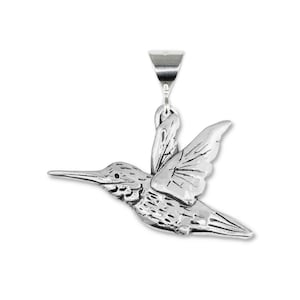 Peut inclure: Pendentif argenté en forme de colibri aux ailes déployées, avec des détails de plumes et un long bec. Le pendentif est doté d'une boucle pour une chaîne et se détache sur un fond blanc.