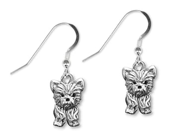 May include: A pair of silver-colored dog-shaped earrings. Each earring features a detailed miniature sculpture of a dog with a textured coat and a small loop for the ear wire. The earrings are suspended from simple, curved ear wires.