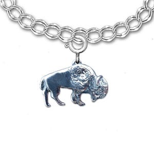 Op de afbeelding: Een zilveren bedelarmband met een buffel hanger. De bedel, die een bizon voorstelt, is bevestigd aan een schakelarmband. De afbeelding staat tegen een witte achtergrond, waardoor de details van de armband worden benadrukt.