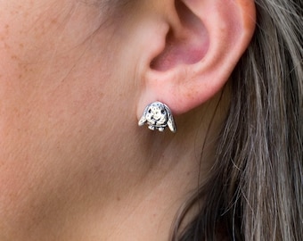 May include: A silver-colored bunny stud earring is shown on an ear. The earring is a detailed, three-dimensional design of a bunny with its ears down. The bunny's face has small, dark spots, and the earring is attached to the earlobe.