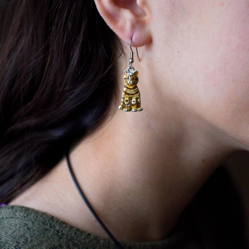 May include: Silver dangle earrings with a gold cat charm. The cat is sitting with its tail curled around its legs.