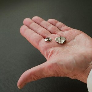 Puede incluir: Dos broches de coraz&oacute;n plateados con cierre de pin. Los broches se muestran en una mano, con la parte posterior de los broches mirando hacia la c&aacute;mara.