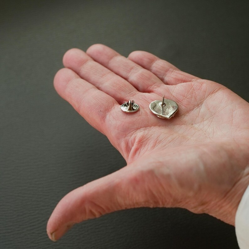 Puede incluir: Dos broches de coraz&oacute;n plateados con cierre de pin. Los broches se muestran en una mano, con la parte posterior de los broches mirando hacia la c&aacute;mara.