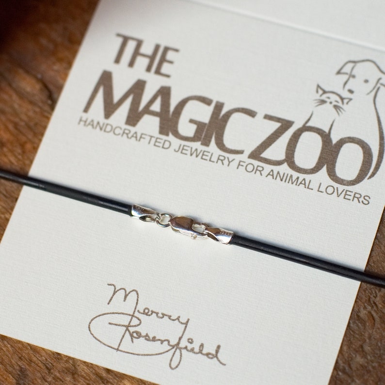 May include: A black leather cord necklace with a silver clasp. The necklace is on a white card with the text "The Magic Zoo Handcrafted Jewellery for Animal Lovers".