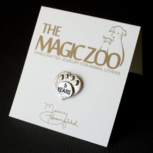 Puede incluir: Encanto plateado en forma de coraz&oacute;n con una huella de pata y el texto "5 Years" en una tarjeta blanca con el texto "The Magic Zoo Joyer&iacute;a artesanal para amantes de los animales" en letras doradas.