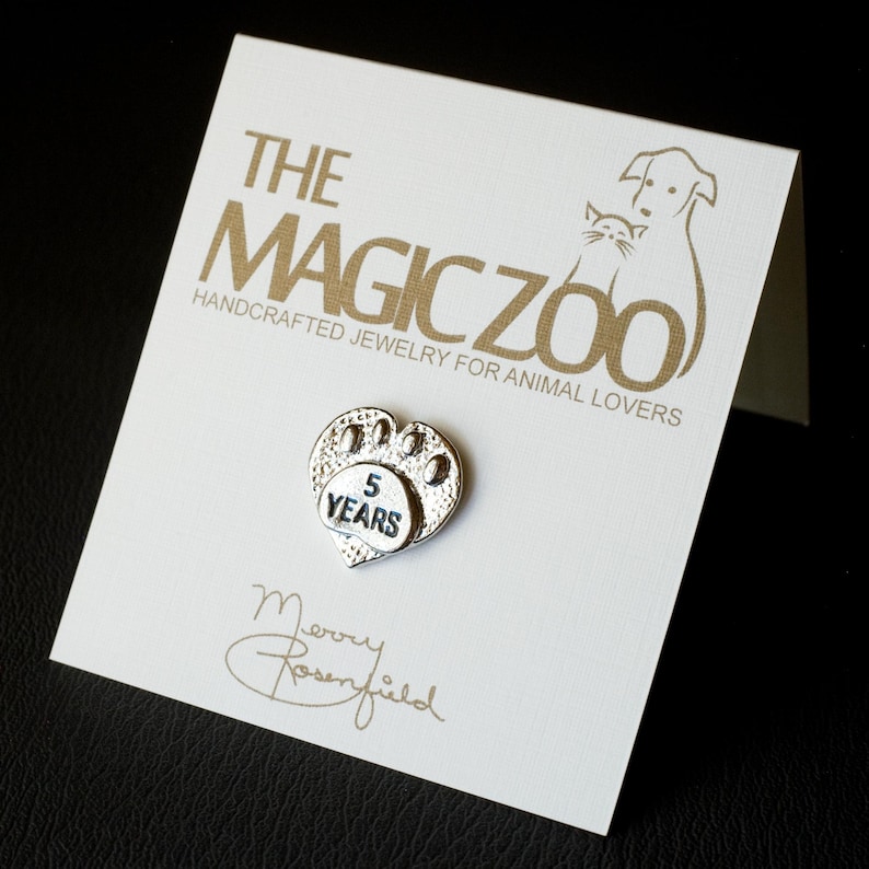 Puede incluir: Encanto plateado en forma de coraz&oacute;n con una huella de pata y el texto "5 Years" en una tarjeta blanca con el texto "The Magic Zoo Joyer&iacute;a artesanal para amantes de los animales" en letras doradas.