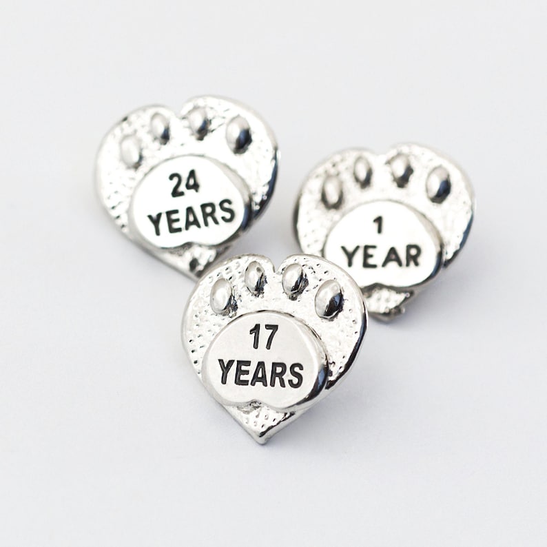 Puede incluir: Tres dijes de plata en forma de coraz&oacute;n con huellas de patas y las palabras "24 A&Ntilde;OS", "1 A&Ntilde;O" y "17 A&Ntilde;OS" grabadas en ellos.