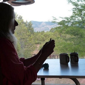 May include: A person sits at a table with two mugs, looking out of a window at a view of trees and mountains.