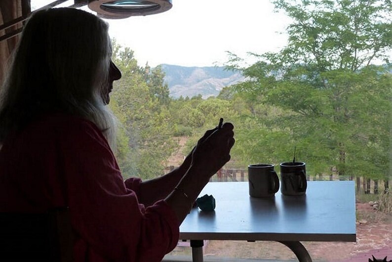 May include: A person sits at a table with two mugs, looking out of a window at a view of trees and mountains.