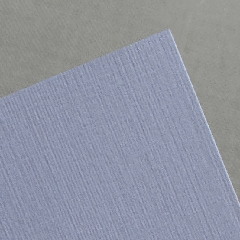 May include: A close-up of a light blue textured paper with a subtle grain pattern. The paper is slightly angled, revealing a gray textured surface underneath.