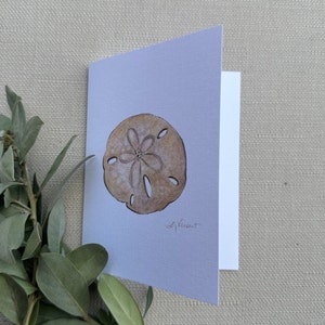 May include: A watercolor painting of a sand dollar on a light blue background. The sand dollar is brown and has a five-pointed star in the center. The artist's signature, "S. Vancut", is in the bottom right corner.
