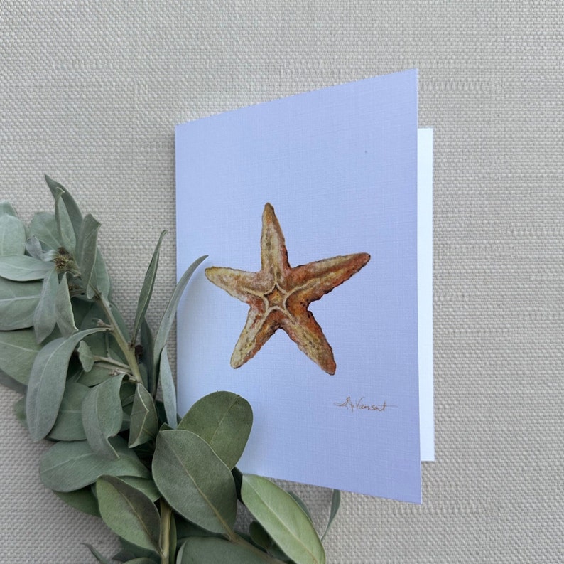 May include: A watercolor painting of a starfish on a light blue card. The starfish is a light brown color with orange accents. The artist's signature is in the bottom right corner.