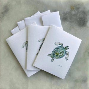 May include: A stack of white greeting cards with matching envelopes. Each card features a watercolor illustration of a sea turtle in shades of green and blue. The cards have a textured surface.