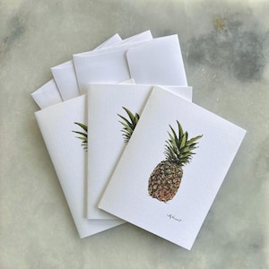 Könnte beinhalten: Ein Set weißer Grußkarten mit passenden Umschlägen. Jede Karte zeigt eine Aquarellillustration einer Ananas mit grünen Blättern und einem braunen Körper. Die Karten sind fächerförmig angeordnet.