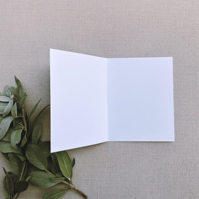 May include: A blank white card with a folded edge, sitting on a light beige textured surface. A sprig of green leaves is in the lower left corner of the image.