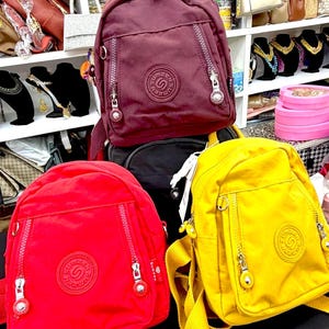 May include: Three small nylon backpacks in burgundy, red, and yellow. Each bag features a top handle, adjustable shoulder straps, two vertical zippered front pockets, and a circular logo patch on the front with the text Sumzen in the center.
