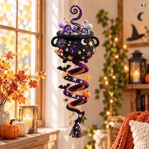 May include: A crocheted hanging decoration featuring a black cauldron filled with purple and green bubbles, topped with a swirling smoke effect. A long, spiral tail extends downward, adorned with small crocheted bats, stars, and pumpkins.