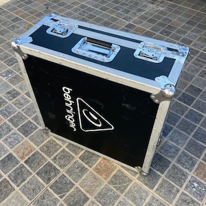 Puede incluir: Un estuche de transporte rectangular negro con bordes de metal plateado y esquinas reforzadas. Cuenta con un asa central, dos cierres de seguridad y el logotipo blanco de Behringer impreso en el lateral. Es una unidad de almacenamiento.