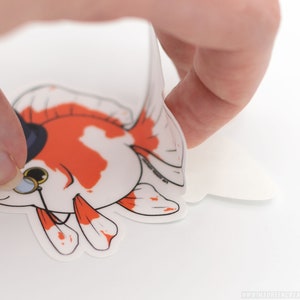 Puede incluir: Una pegatina blanca con un pez dorado de dibujos animados que lleva un sombrero de copa y un mon&oacute;culo. El pez dorado es naranja y blanco y tiene un ojo negro. La pegatina se est&aacute; despegando de un respaldo blanco.