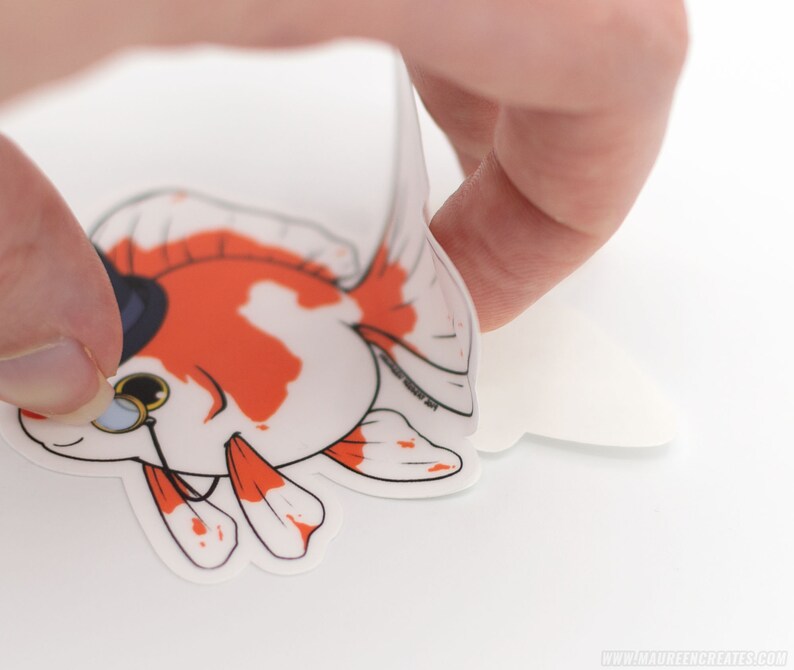Puede incluir: Una pegatina blanca con un pez dorado de dibujos animados que lleva un sombrero de copa y un mon&oacute;culo. El pez dorado es naranja y blanco y tiene un ojo negro. La pegatina se est&aacute; despegando de un respaldo blanco.