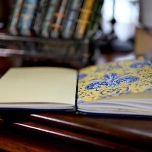 May include: An open notebook with a plain cream-colored page on the left and a decorative page on the right featuring a yellow background with a detailed blue floral and scrollwork pattern. The notebook is bound with visible stitching.
