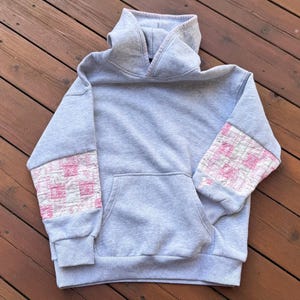 May include: A light gray hooded sweatshirt featuring a front kangaroo pocket and long sleeves with quilted patchwork panels in shades of pink and white. The garment has a high neckline and ribbed cuffs at the wrists and hem.