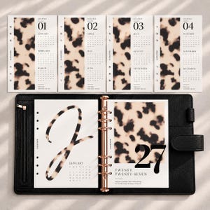 May include: A black leather ring-bound planner containing calendar inserts for the year 2027. The pages feature a beige and black tortoiseshell pattern, with clear black typography displaying months, dates, and the year twenty twenty-seven.
