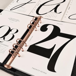 May include: A black leather binder with rose gold rings holding white calendar pages. The pages feature large black typography for the number 27 and the text TWENTY-SEVEN, along with smaller monthly calendars for July, August, and September.