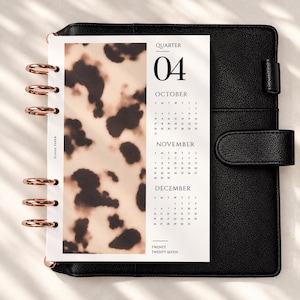 May include: A black leather planner with rose gold disc binding rings holding a calendar insert. The insert features a beige and brown tortoiseshell pattern on the left and the text QUARTER 04, OCTOBER, NOVEMBER, DECEMBER, and TWENTY TWENTY-SEVEN.