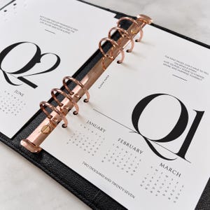 May include: A black leather planner with rose gold rings featuring white pages for the year two thousand and twenty-seven. The pages display Q1 and Q2 calendars for January through June, with motivational text printed in black ink on the white paper.