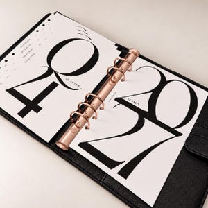 May include: A black leather binder with a rose gold six-ring mechanism holding white pages. The left page features the number 40, the text QUARTER, and the words PLAN DEEPER, BUILD SOFTER, CREATE MORE, ELAVAE PAPER. The right page displays 2027 and SEASON.