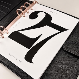 May include: A black leather binder with rose gold rings holding a white page featuring the large black number 27. The page includes the text ELAVAE PAPER, TWENTY, and TWENTY-SEVEN printed in a small black serif font on the white background.