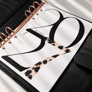 May include: A black leather planner with rose gold rings holding a white page. The page features large black typography reading 2024, with the word SEASON printed in small black letters. Part of the number 4 has a tan and black leopard print pattern.