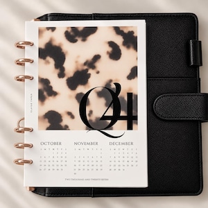 May include: A planner insert featuring a beige and black tortoiseshell pattern with the large black text Q4. Below, a calendar displays October, November, and December for the year two thousand and twenty-seven. The page has rose gold metal discs.