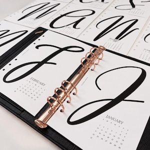 May include: A black leather binder with a rose gold six-ring mechanism holding white calendar pages. Each page features a large black handwritten script letter for the month, including January and February, with a small monthly grid below.