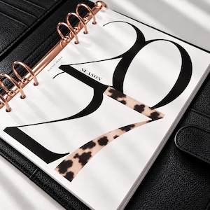 May include: A white planner page with large black numbers 2024, where the number 4 features a tan and black leopard print pattern. The text ELAVAE PAPER and SEASON are printed in black. The page is held by rose gold rings in a black binder.
